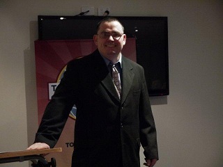 picture of David in a suit standing next to a lectern, speaking about his book and living with a brain injury